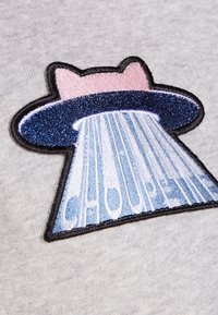 Naszywka z haftem przedstawiająca kreskówkowe UFO z różowym rondem i niebieskimi błyszczącymi detalami, emitujące światło, z napisem "CHOUPETTE" poniżej w jasnoniebieskim kolorze.