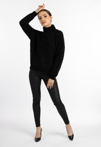 Schwarzer, flauschiger Rollkragenpullover mit lockerer Passform, kombiniert mit glänzenden, schwarzen, figurbetonten Leggings und schwarzen High Heels, vor einem weißen Hintergrund posiert.