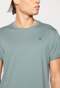 Camiseta corta de manga verde claro hecha de algodón, con un cuello redondo y un pequeño logo impreso en un verde más oscuro en el pecho.