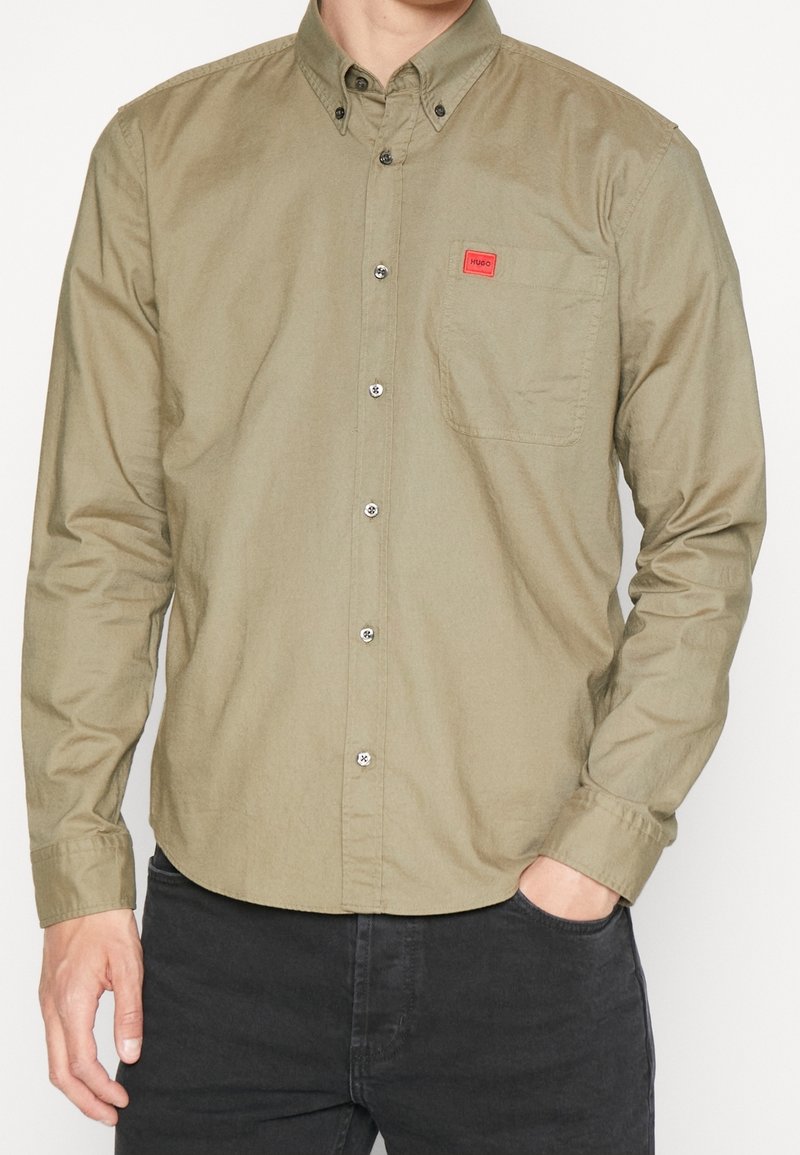 Homme portant une chemise olive à manches longues boutonnée avec poche poitrine et écusson logo Hugo rouge, associée à un pantalon noir, main dans la poche.