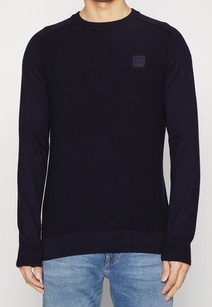 Mann trägt einen enganliegenden schwarzen Strickpullover mit langen Ärmeln und einem kleinen quadratischen Logo auf der Brust, kombiniert mit hellblauen Jeans.