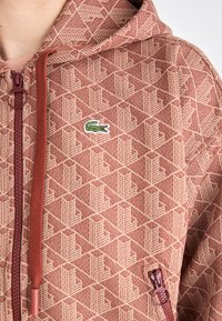 Hettejakke med glidelås i et rødt og beige geometrisk mønster, utstyrt med et logo-patch og kontrasterende glidelåsdetaljer. Myk tekstur og avslappet passform.