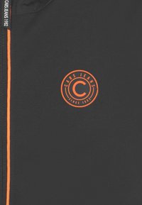 Černá látka s oranžovým zipem a oranžovým kruhovým logem s nápisem "CARS JEANS SINCE 1982" blízko středu.