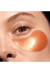 Hydrating eye patch in a shiny, orange-gold color applied under a brown eye, alongside soft, smooth skin and natural eyelashes.