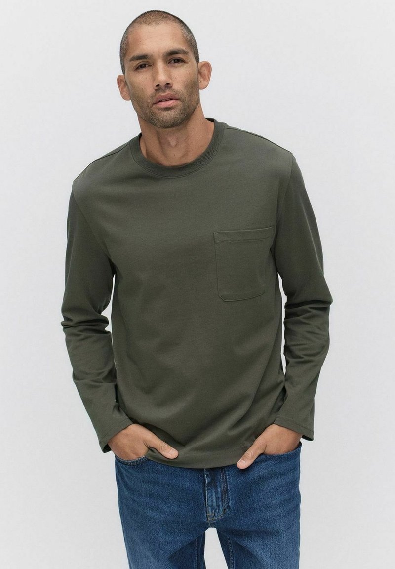 Långärmad skjorta i mörk olivgrön, med rundad halsringning och bröstficka, tillsammans med blå denimjeans. Mjuk tygtextur.