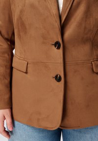 Blazer en suède marron avec deux boutons noirs, coupe ajustée et poches latérales, présentant une texture lisse et des lignes épurées.