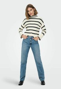 Balts un melns svītrains augstas apkakles džemperis ar platām piedurknēm, kopā ar zilām taisnkājām džinsa biksēm un melniem potīšu zābakiem.