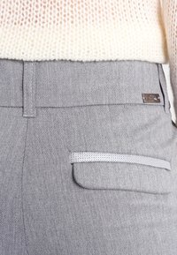 Šedé kalhoty v herringbone vzoru s elegantním designem, jemnou strukturou a malou kapsou s dekorativním světle šedým akcentem a kovovým hardwarem.