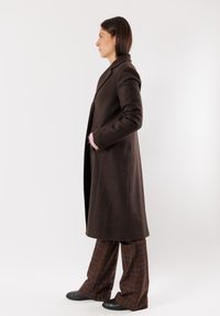 Cappotto lungo in lana marrone con silhouette aderente e revers a punta. Presenta tasche laterali e dettagli in maglia rosa ai polsini.