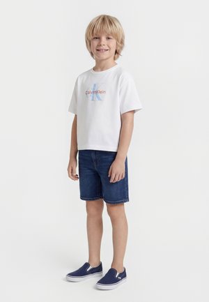 Ξανθός αγόρι όρθιο και χαμογελαστό, φοράει λευκό μπλουζάκι Calvin Klein, τζιν σορτς και μπλε ναυτικό παπούτσια slip-on, σε λευκό φόντο.