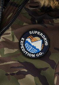 Tarnjackenstoff mit einem runden Patch, das blaue Berge und die Sonne zeigt, mit gesticktem Text "SUPERDRY EXPEDITION CO." in weißem Rand.