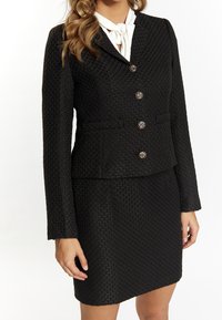 Completo nero con texture, include un blazer aderente caratterizzato da bottoni decorativi e una gonna abbinata, indossato con una camicetta bianca con fiocco.