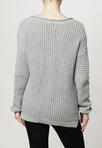 Femme portant un pull épais en tricot gris clair, vue de dos, associé à un pantalon noir sur un fond neutre.