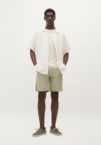 Cămașă ușoară din in cu mânecă scurtă, de culoare albă, suprapusă peste un tricou crem, asociată cu pantaloni scurți de culoare verde măslin și pantofi verzi cu tălpi bej.