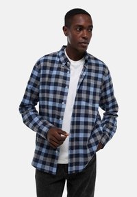 Chemise à carreaux bleu, marine et gris. Matière en coton, manches longues, léger détail de poche, portée sur un t-shirt blanc.