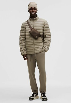 Beige gesteppte Jacke mit Reißverschluss, beige Jogginghose, dunkle Turnschuhe und eine braune Umhängetasche. Texturiertes Material, praktisches Schichtdesign.