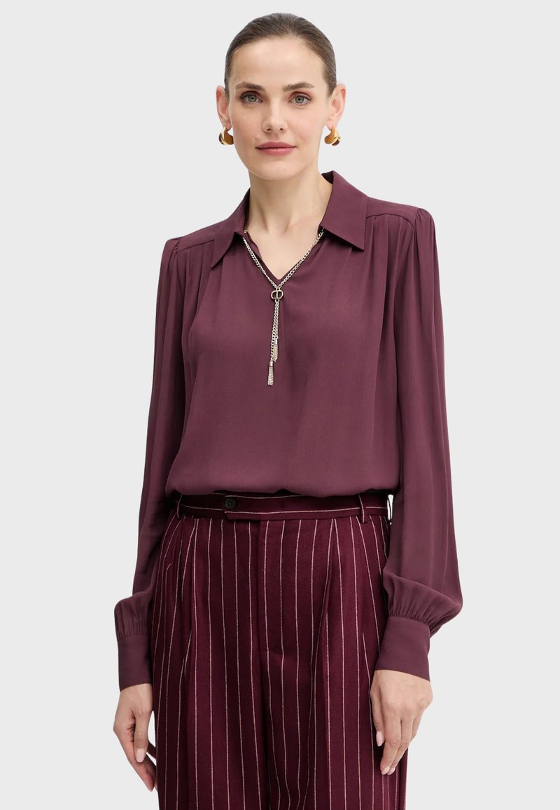 Donna che indossa una blusa borgogna con maniche lunghe a palloncino e pantaloni a righe a vita alta, accessoriate con orecchini a cerchio dorati e una collana d'argento.