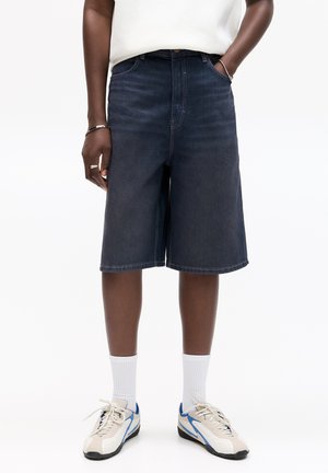 Shorts en denim bleu foncé à coupe droite, avec poches latérales et taille mi-haute, associés à des baskets beige et bleu et des chaussettes blanches.