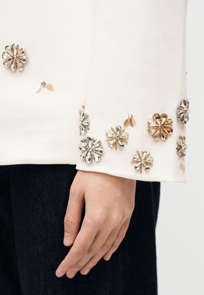 Χέρι που ακουμπά στο πλάι, φορούσε λευκό μπουφάν με διακοσμητικά λουλούδια από πέτρες στο μανίκι και ορατό μαύρο παντελόνι.