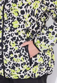 Mano in tasca di una giacca bianca con stampa leopardata nera e verde neon, zip nera, polsini elastici e etichetta HYBRAR sulla manica.