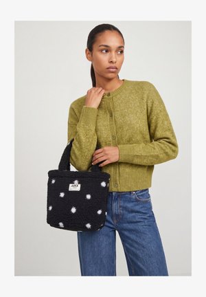 Jeune femme en pull vert boutonné et jean bleu tient un sac à main noir duveteux à pois blancs avec une étiquette à l'avant.
