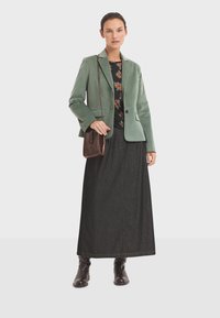 Woman wearing green blazer, floral black top, long dark skirt, brown boots, and carrying a small brown crossbody bag, standing against white background.