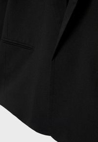 Nærbillede af sort blazerstof, der viser pouch-detalje og skarp reverskant mod en ensfarvet baggrund.