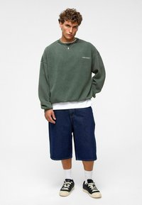 Pull vert surdimensionné avec épaules tombantes, associé à un short en denim noir arrivant au mi-mollet, et des baskets noires avec accents blancs.