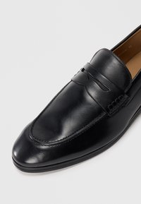 Mocassin en cuir noir au design épuré, mettant en avant des détails de couture et une texture lisse. Bout arrondi et accent subtil sur le dessus.