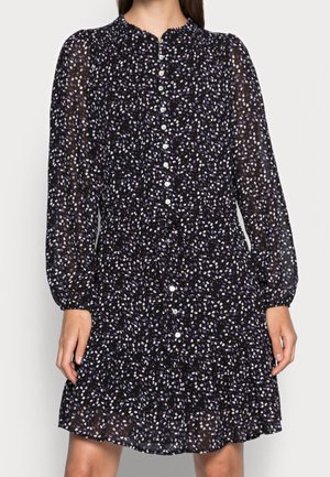 Vrouw draagt een zwarte jurk met lange mouwen, kleine witte en paarse bloemenpatronen en knopen aan de voorkant, staand tegen een effen achtergrond.