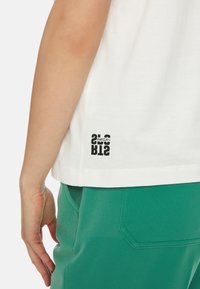 Weißes Baumwoll-T-Shirt mit einem schwarzen Grafik-Logo auf der unteren linken Seite, kombiniert mit grünen Stoffhosen, die eine kontrastierende Textur aufweisen.