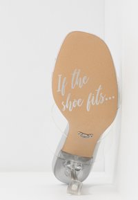 Semelle transparente d'une chaussure à talon haut avec un texte argenté indiquant "If the shoe fits…" et une bride ainsi qu'un talon transparents.