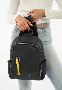 Svart texturerad ryggsäck med två svarta remmar, silverfärgade dragkedjor och en gul accent på dragkedjehandtaget. Har en kompakt, rund form.