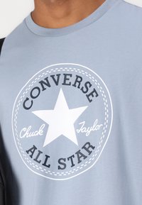 Tricou bleu deschis cu un grafic circular alb, având o stea neagră și textul "CONVERSE ALL STAR", realizat dintr-un material moale din bumbac.