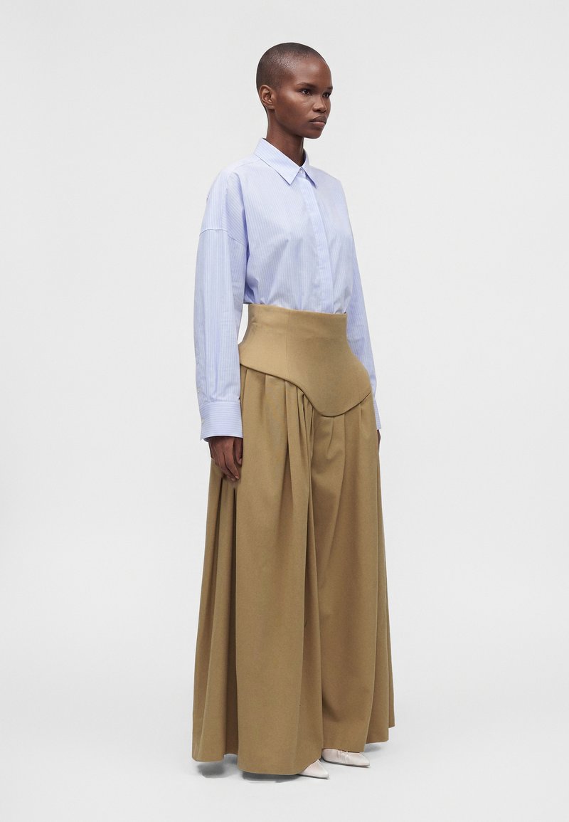 Femme portant une chemise bleu clair boutonnée rentrée dans un pantalon large beige taille haute avec un panneau structuré à la taille, debout devant un fond uni.