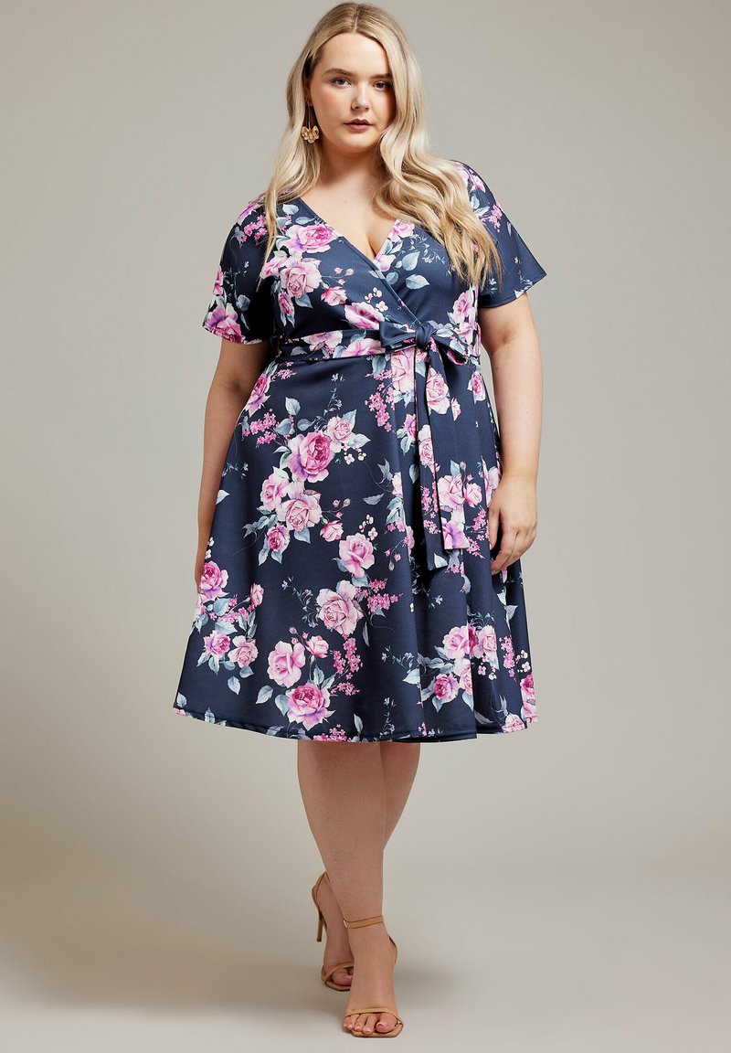 Vestido envolvente floral en tejido azul marino con mangas cortas, un lazo en la cintura y rosas rosas con hojas verdes. Textura suave, hasta la rodilla.