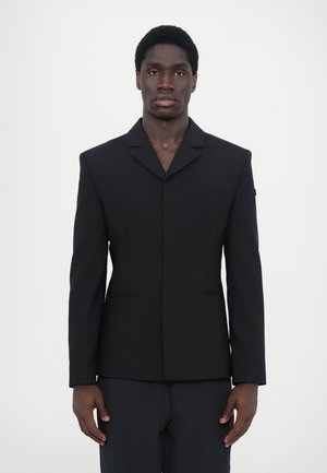 Schwarze maßgeschneiderte Sakkojacke mit fallendem Revers, einer Tasche und strukturierten Schultern. Glattes Material und klare Linien; minimalistisches Design.