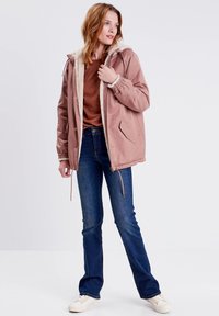 Rosa parkas jacka med fleecefodrad huva, dragsko i midjan och sidofickor, tillsammans med en brun topp och blå jeans. Lätta skor.