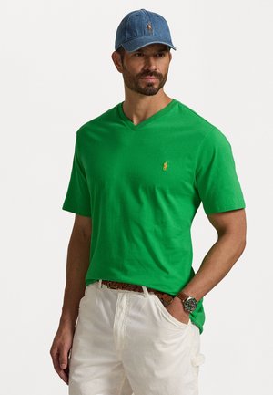 Green v-neck t-shirt in cotton, featuring a small yellow embroidered logo on the chest, paired with a light blue denim cap. White shorts visible.