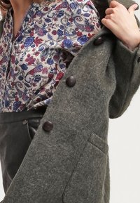 Grå ullkappa med stora bruna knappar, bärs över en blommönstrad blus med blå och röda blommor på en ljus bakgrund.