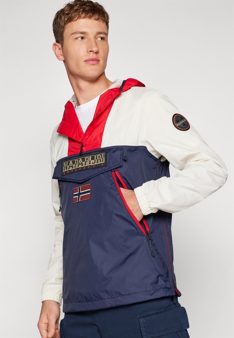 Giovane uomo che indossa una giacca a blocchi di colore con maniche bianche, dettagli rossi, corpo blu navy e loghi del marchio sul petto e sulla manica.