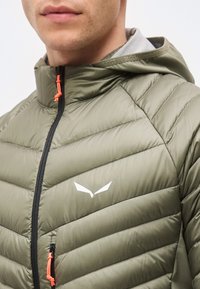 Άνδρας που φοράει μπουφάν χακί με κουκούλα, μαύρο φερμουάρ και πορτοκαλί τσερτζελάκια φερμουάρ, με λευκό φτερωτό λογότυπο στο στήθος.