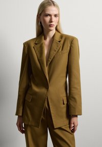 Blazer ajustado de color verde oliva con solapas afiladas, cierre de un solo botón, dos bolsillos y una tela de textura suave. Lleva pantalones a juego.