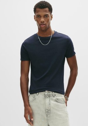 Mand iført mørkeblå kortærmet T-shirt, lyseblå jeans, sølvkæde og armbånd, står mod en ensfarvet hvid baggrund.