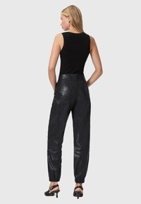 Vrouw met blond haar draagt een mouwloos zwart topje, zwarte hoge leren joggingbroek met zakken en zwarte hoge sandalen, terwijl ze met haar rug naar de camera staat.