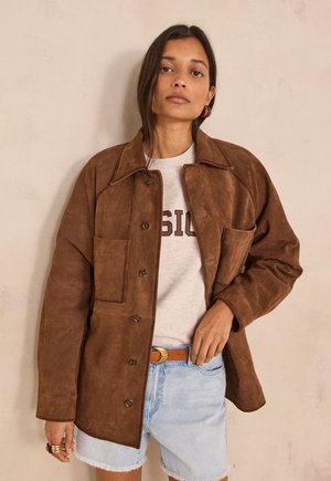 Femme portant une veste en daim marron à boutons, un t-shirt graphique beige, un short en jean clair et une ceinture marron, debout devant un fond uni.