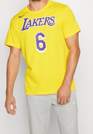 Homme portant un t-shirt jaune des Lakers numéro 6 et un pantalon de survêtement Nike gris clair, debout avec la main droite détendue et la main gauche près du pantalon.