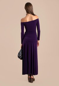 Robe violette décolettée avec des manches longues, détails froncés à la taille, jupe fluide, portée avec un sac à main noir texturé et des chaussures à plateforme.