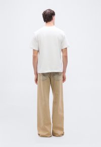 Άνδρας με πλάτη φοράει απλό λευκό κοντομάνικο μπλουζάκι και φαρδιά μπεζ παντελόνια σε απλό ανοιχτό φόντο.