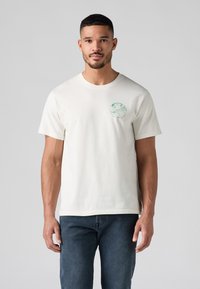 Homme portant un t-shirt blanc à manches courtes avec un graphique de globe vert et du texte sur le côté gauche de la poitrine, associé à un jean bleu marine.